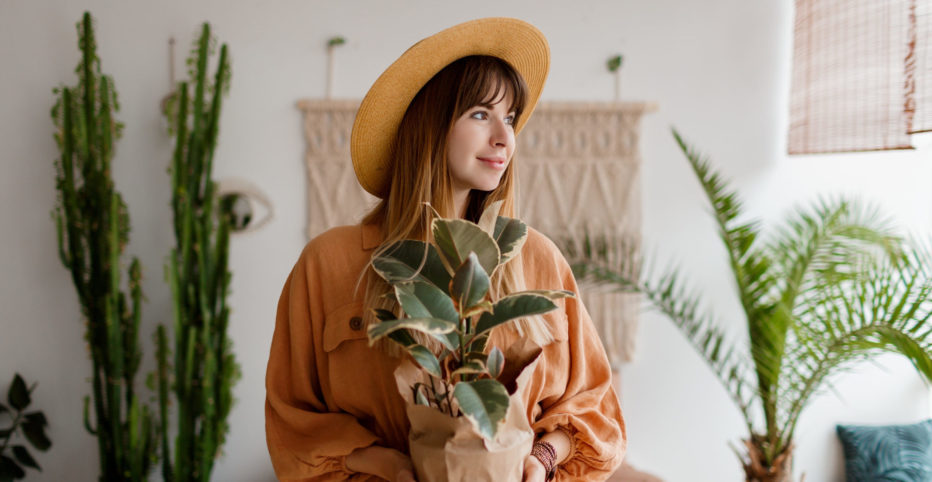 Woman in straw hat and linen dress holding home plant over stylish interior boho background