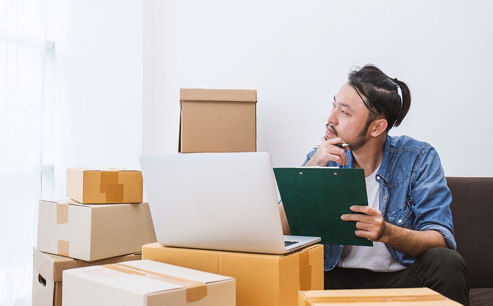 Asian man works at home on his laptop surrounded by moving boxes