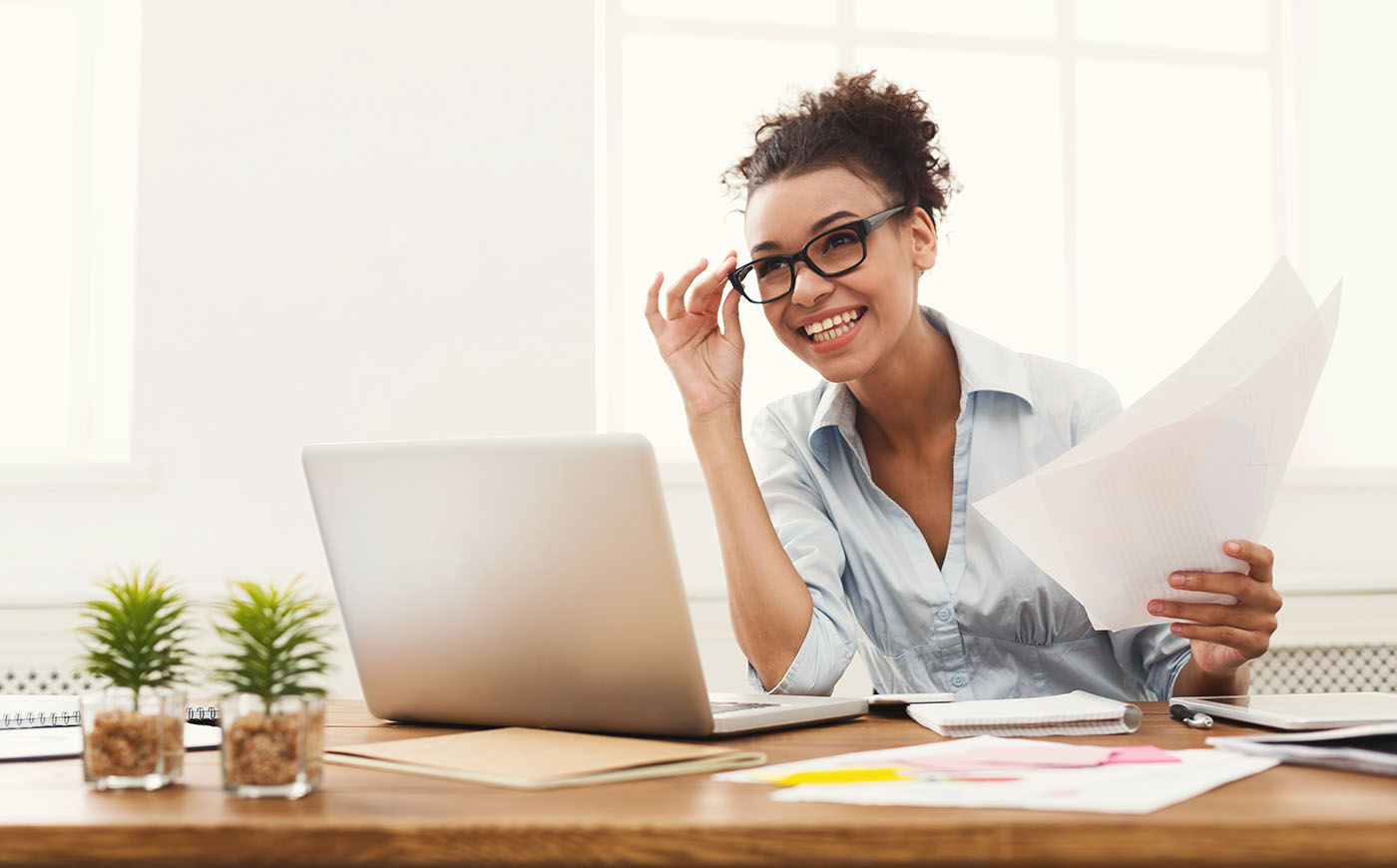 Happy smiling Black business woman in formal wear sitting at wooden desk in modern office and reading report document