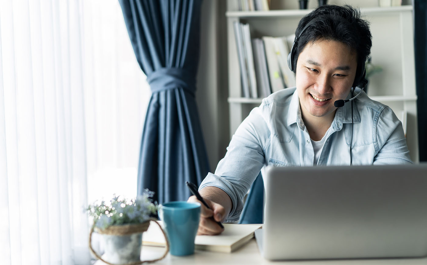 Asian male at home on computer while talking virtually and taking notes