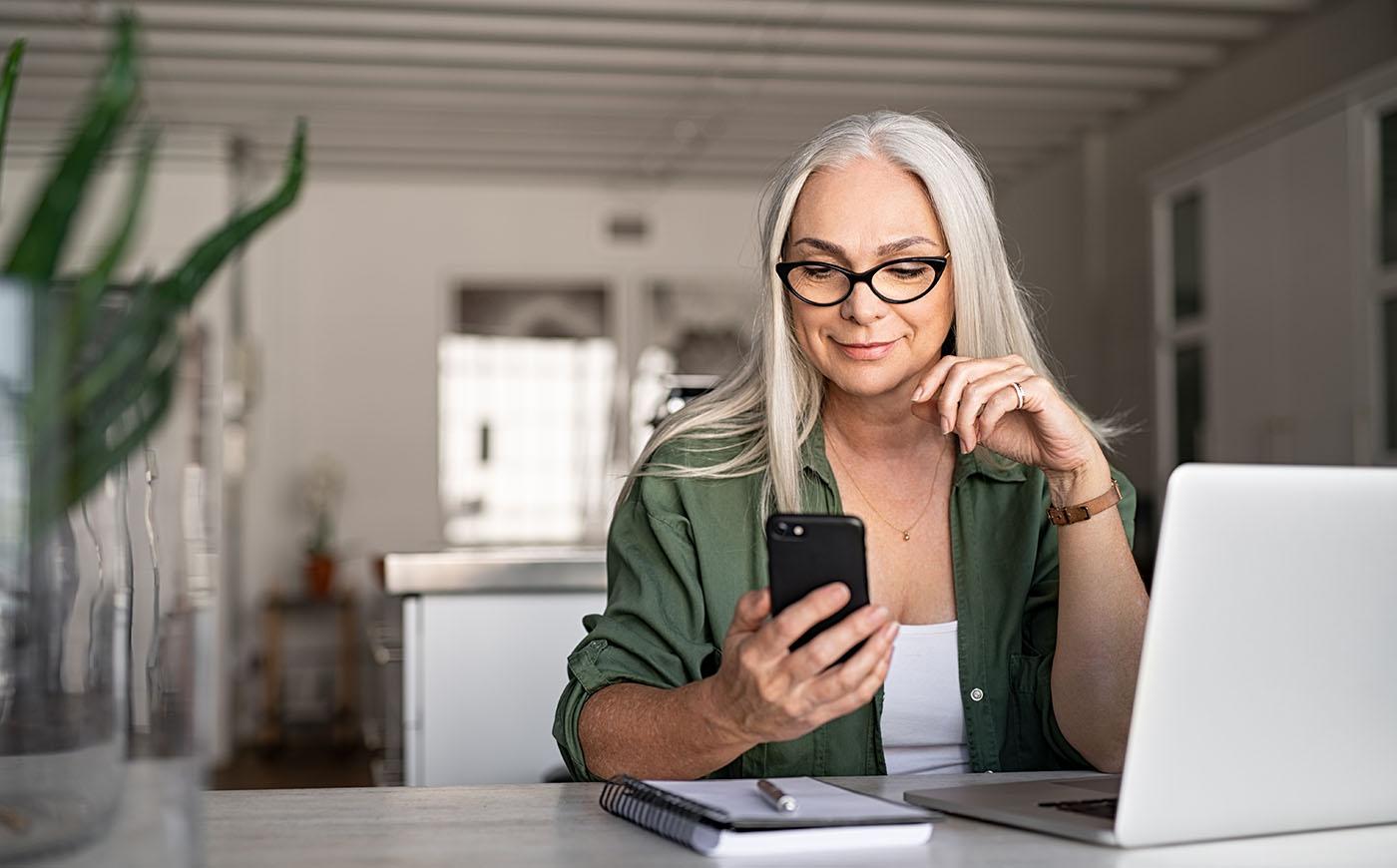 Older woman with long gray hair smiles at her phone as she sits in front of her laptop in her kitchen
