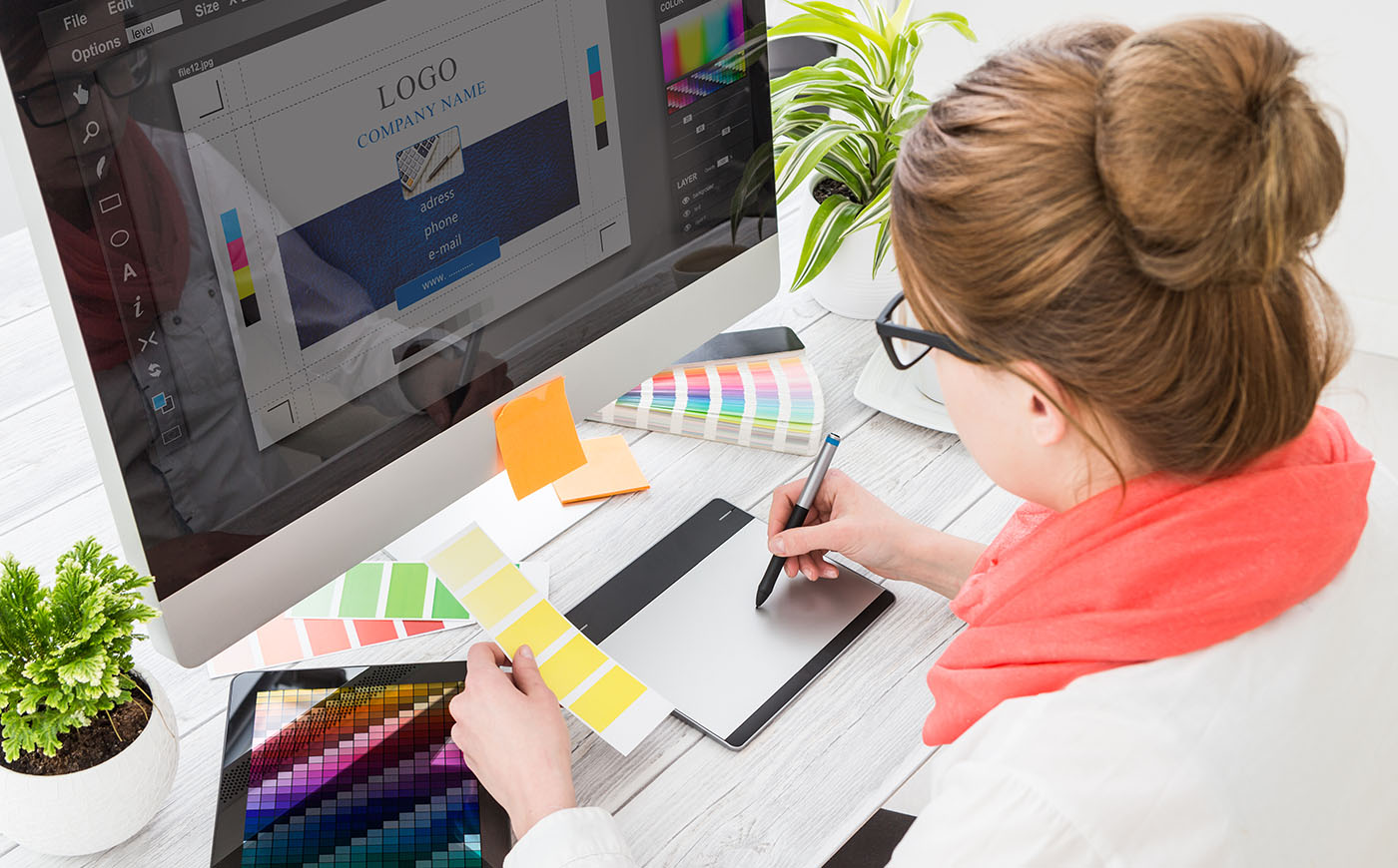 Woman consults digital and printed color palettes as she works on a logo design on her desktop computer using a tablet and pen