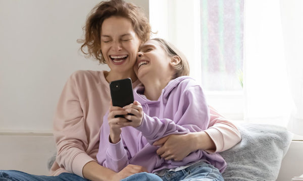 Mother and daughter joyously laugh together at home in bed while daughter holds smartphone
