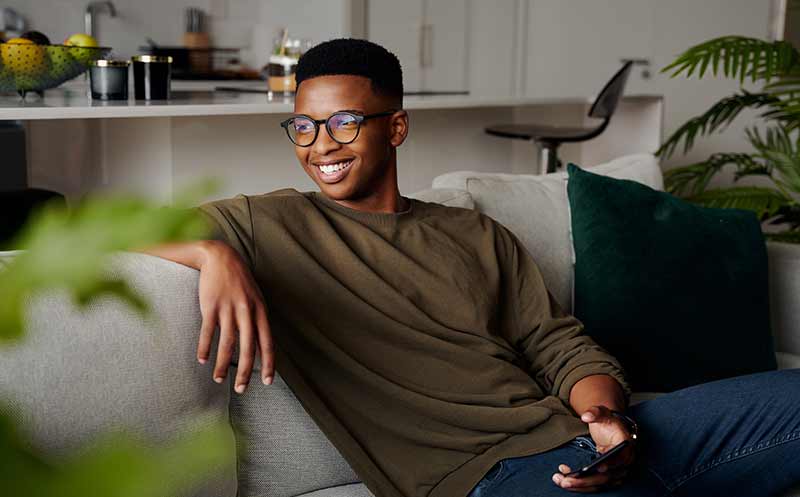 Young adult Black man cheerfully relaxing on sofa holding smartphone in modern apartment