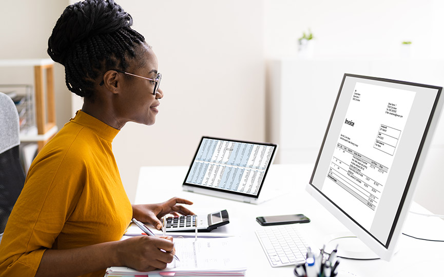 Black professional woman sitting at her desk at home as she calculates numbers on her desktop computer and takes notes on paper