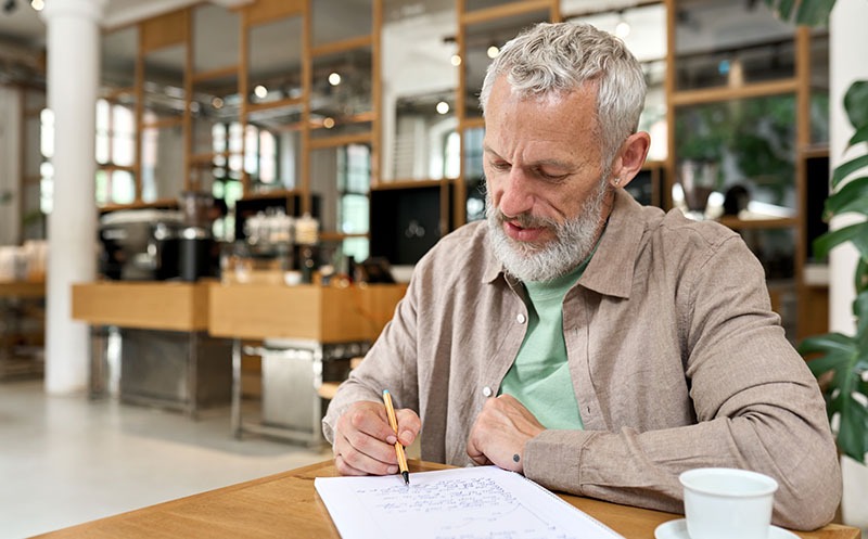 Older man writing his thoughts in a notebook at a table in a cafe