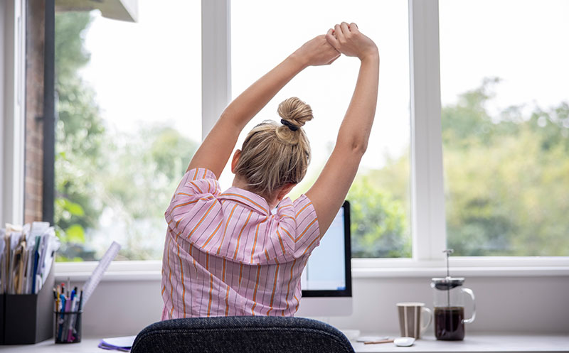 Rear view of woman working from home on computer in home office stretching at desk in front of large window
