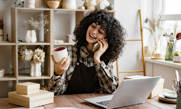 Woman potter entrepreneur talking to buyer of handmade mug sitting at table and taking order using laptop in her workshop