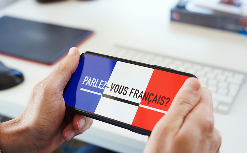 Person sitting at a desk with a language-learning app open on their smartphone that says "do you speak French?" written in French on the phone