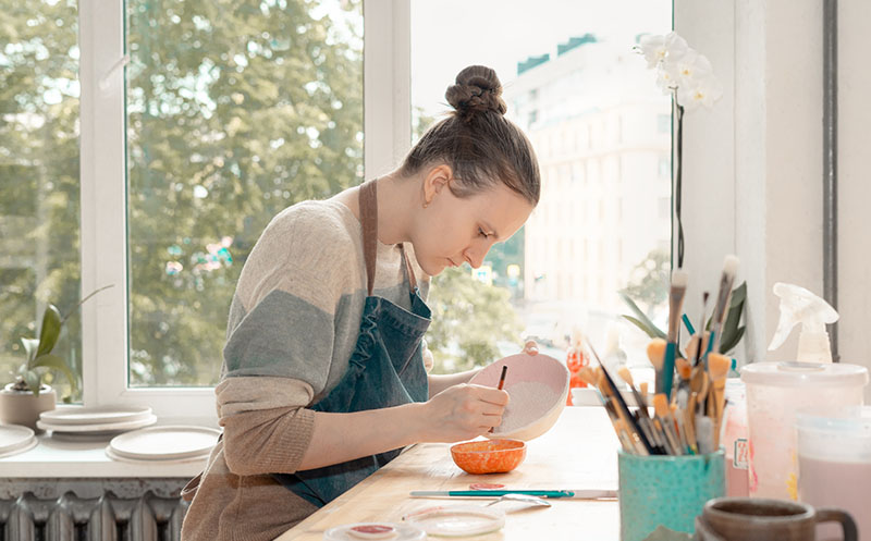 Skilled young woman in apron sitting at table and drawing on ceramic bowl in pottery workshop