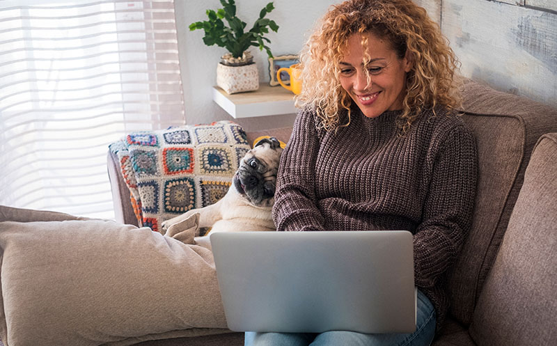 Woman smiles and sits cozy on couch wearing a sweater with a laptop on her lap and pug snuggled against her side