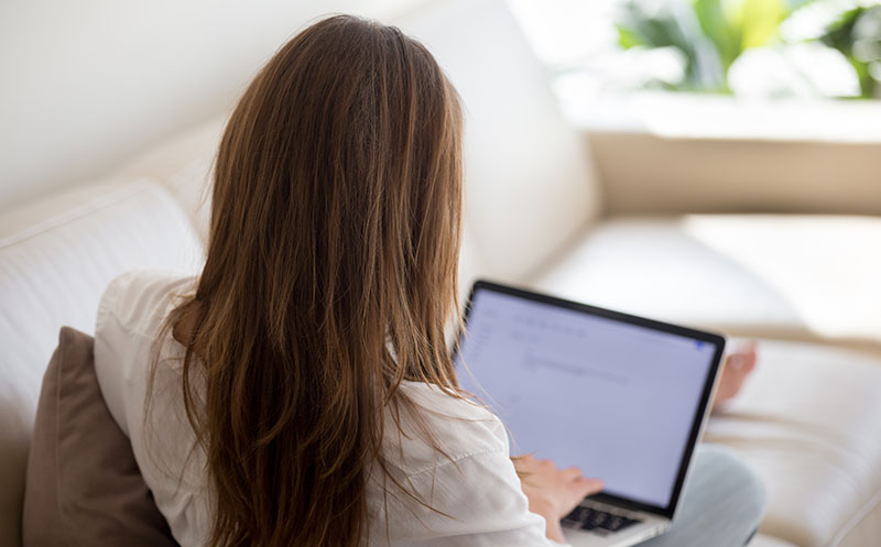Behind shot of a woman sitting on her couch typing an application for a remote job online