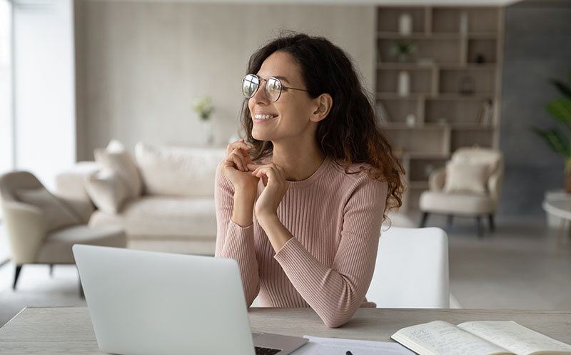 Young woman sitting at her desk at home and optimistically looking out into the distance with a smile