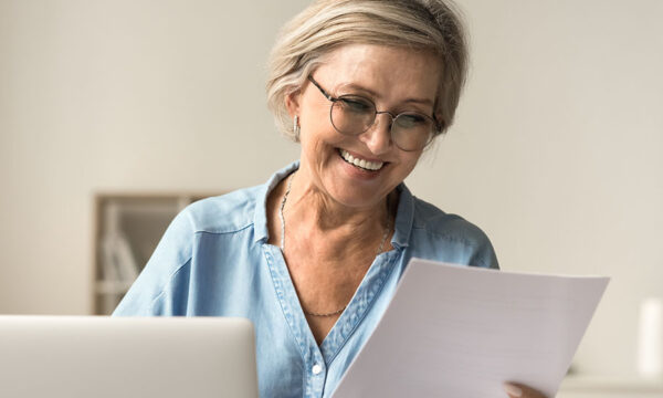 Mature retired accountant smiles at a piece of paper in her hand and her laptop in her home office