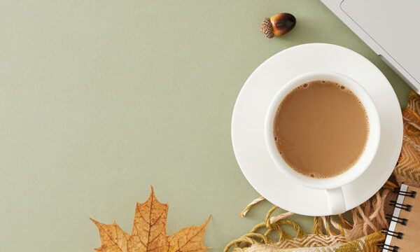 Overhead view of fall-decorated work table that includes a mug of coffee, a laptop, a notebook, pencil, and fall leaves.