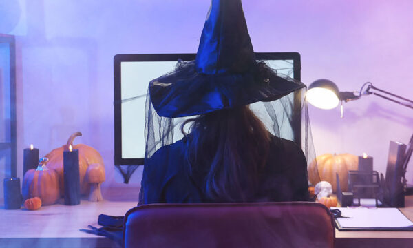 Woman dressed as a witch sitting at a desktop computer at home. The room is lit with purple lights, the desk has pumpkins on it, and a fog is in the air.