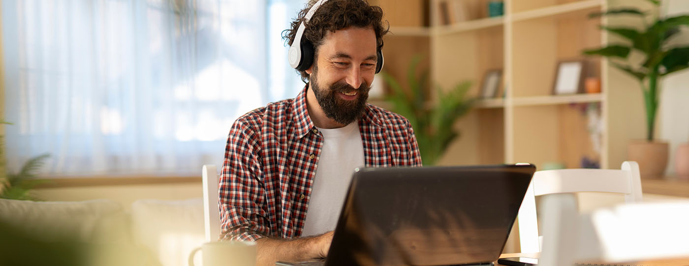 Young man wearing headphones and smiling while working on a laptop at a bright home office